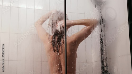 Young woman washes her hair in the shower in the bathroom. A woman washes her long hair with shampoo. A carefree girl takes a long hot shower, washes her hair in a modern bathroom