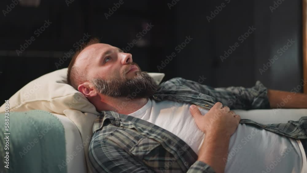 Bearded man with acute heart pain lies on sofa putting hand on chest. Male with high temple waits for relief after taking medications in living room closeup Bearded man with acute heart pain lies on sofa putting hand on chest. Male with high temple waits for relief after taking medications in living room closeup