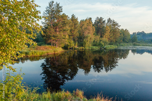 A picturesque reflection of the sky and trees in a forest river. The concept of vacation, summer, fishing, rest on the river bank, calendar, cover. Horizontal photo. 