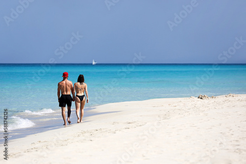 Couple in swimwear walking by white sand beach and holding hands on background of azure ocean waves. Man and woman together, romantic leisure on tropical coast