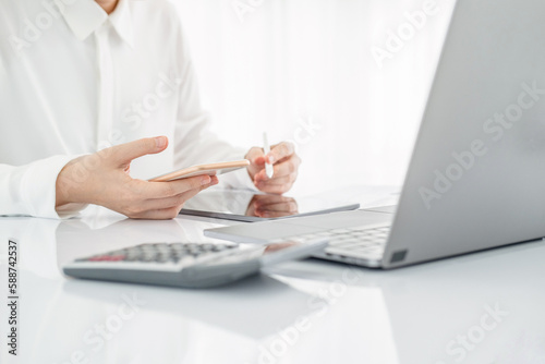 Asian businesswoman in white formal clothes writing note in tablet and checking smart phone. Business woman busy working with documents and tablet  in workstation. close up clean image.