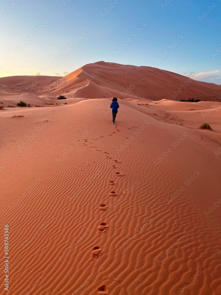 Dunes in the Sahara desert, Merzouga desert, grains of sand forming small waves on the dunes, panoramic view. Setting sun. Morocco. Girl that walk on a sand dune. Shoe prints Dunes in the Sahara desert, Merzouga desert, grains of sand forming small waves on the dunes, panoramic view. Setting sun. Morocco. Girl that walk on a sand dune. Shoe prints