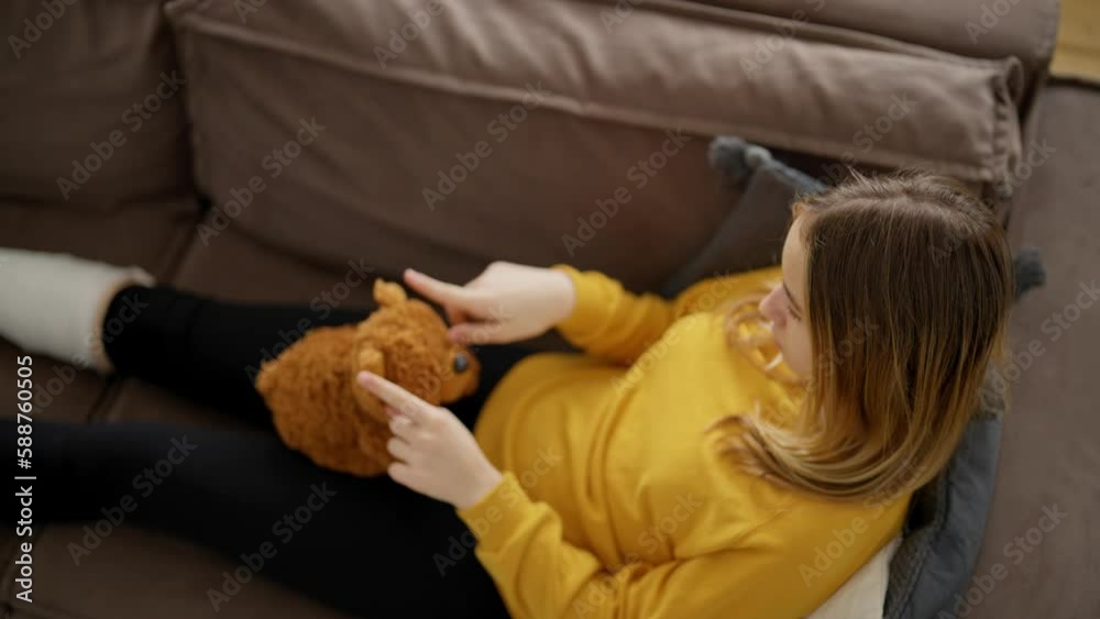 Back view child sitting on couch at home with teddy bear in hands. Girl with broken leg with white plaster cast. Recovery and treatment at home after injury. Support sad children with problem health.