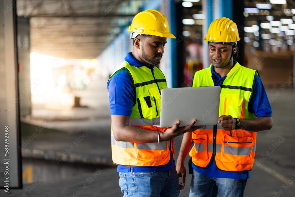 Two men warehouse indian worker wearing safety hardhats helmet working on laptop computer in warehouse of Wholesale Merchandise. shelves, pallets and boxes. Container and warehouse factory