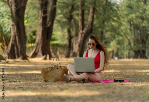 Workcation. Beautiful caucasian white female sexy woman in red bikini swimsuit sitting on the ground relaxingly and using her laptop to work online in a green tropical lush park by the beach photo