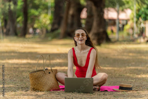 Workcation. Beautiful caucasian white female sexy woman in red bikini swimsuit sitting on the ground relaxingly and using her laptop to work online in a green tropical lush park by the beach photo