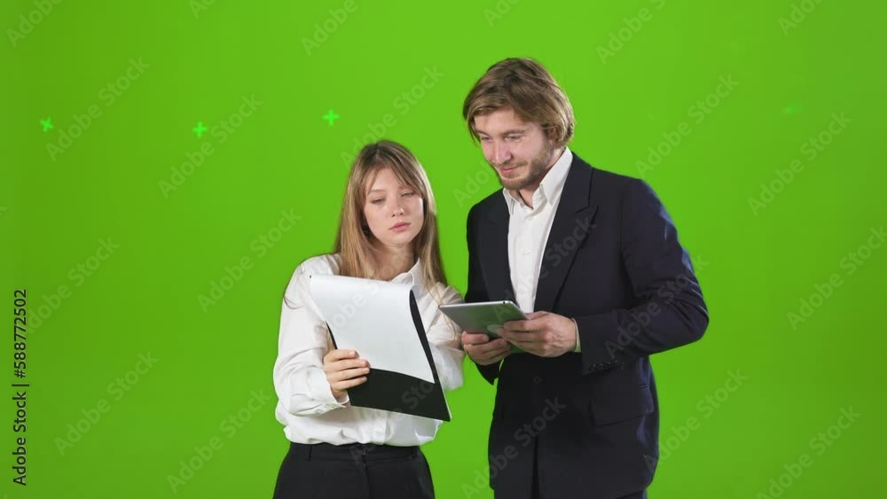 Side view of young business partners standing in studio, working. Attractive female and handsome male holding folder and tablet, discussing. Isolated on green studio background., Side view of young business partners standing in studio, working. Attractive female and handsome male holding folder and tablet, discussing. Isolated on green studio background.,
