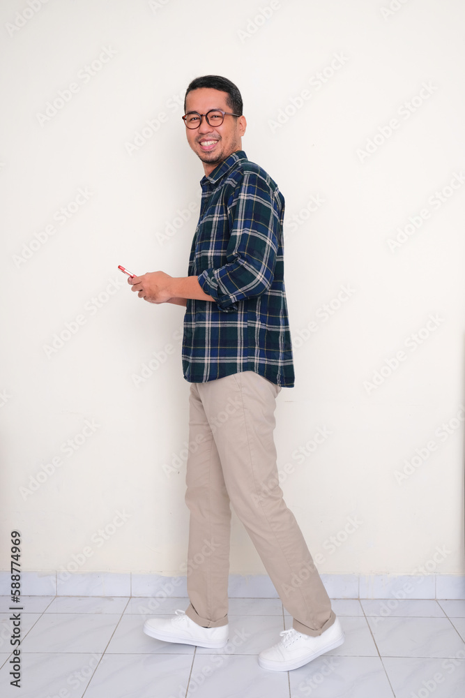 Full length side view photo of Asian man walking and smiling while holding his mobile phone Full length side view photo of Asian man walking and smiling while holding his mobile phone