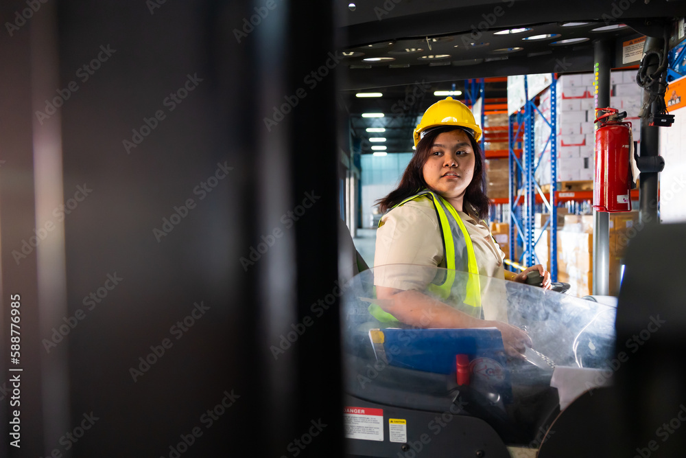 Fototapeta premium Asian fat female warehouse worker drive forklift in warehouse. Warehouse staff worker by goods shelf working in large warehouse factory