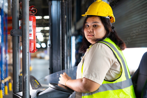 Asian fat female warehouse worker drive forklift in warehouse. Warehouse staff worker by goods shelf working in large warehouse factory