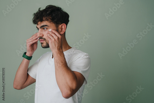 Side view of a man applying face tonic with cotton pad isolated over green wall background with copy space. Skincare morning routine, careful beauty treatment