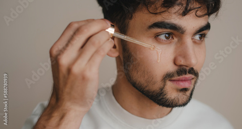 Attractive young man applying essential oil on facial skin while looking away isolated on plain pastel grey background studio. Skin care healthcare cosmetic