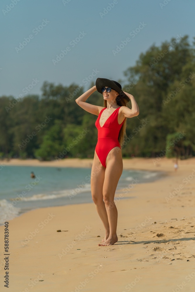 Beautiful sexy caucasian female woman in red one piece swimsuit bikini with black beach hat and sunglasses walking on the beach smile happily enjoying the sea breeze Beautiful sexy caucasian female woman in red one piece swimsuit bikini with black beach hat and sunglasses walking on the beach smile happily enjoying the sea breeze
