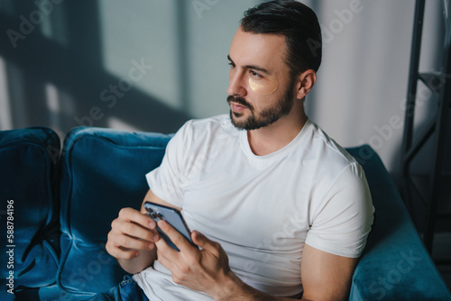 Young serious man using modern smartphone device while sitting on sofa at home, typing an sms message at social network, technology concept. Technology