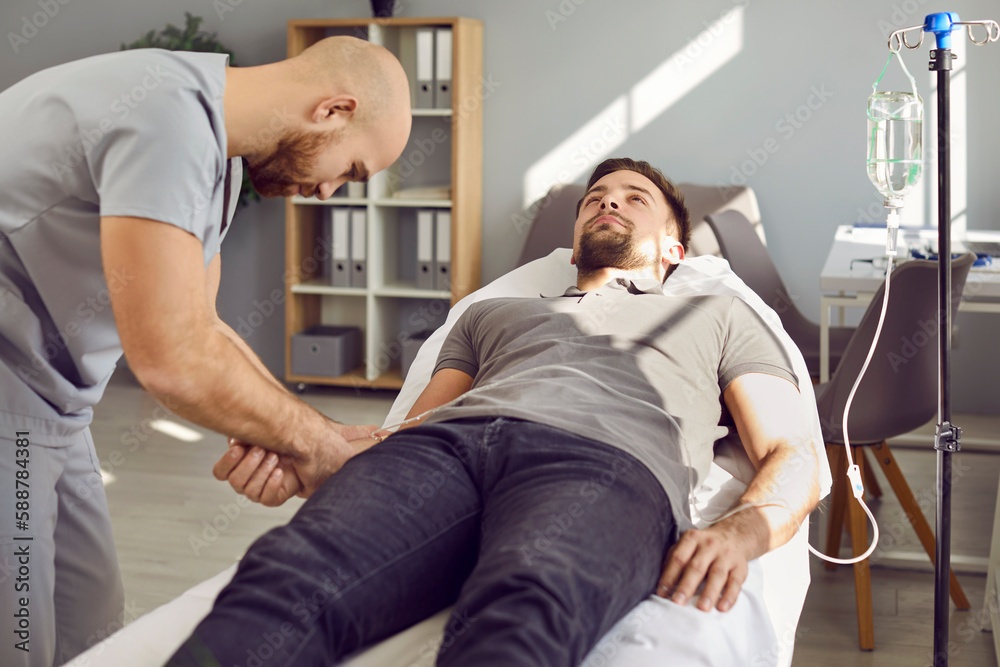 Obraz premium Staff nurse gives intravenous vitamin infusion to young male patient. Professional personnel puts medicine IV line catheter in vein of weak adult man lying on bed in hospital ward or medical office