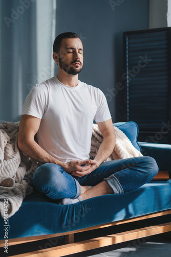 Calm man wearing casual clothes sitting on a sofa in a living room with closed eyes, meditating in the lotus positio. Meditation, relaxation, rest, calmness.