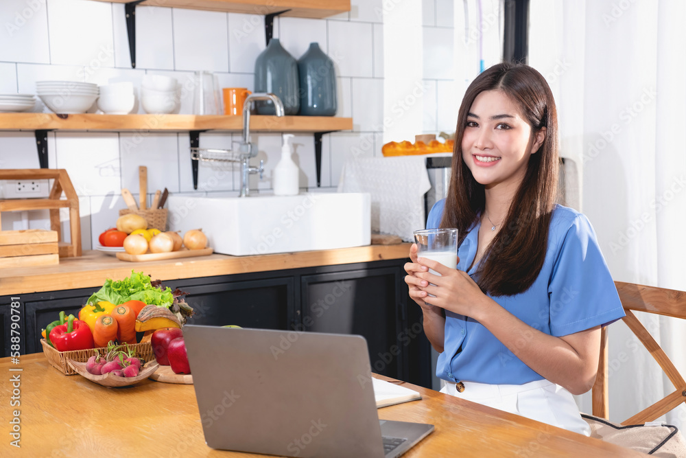 A young woman with a beautiful face in a blue shirt with long hair eating fruit sitting inside the kitchen at home with a laptop and notebook for relaxation, Concept Vacation.