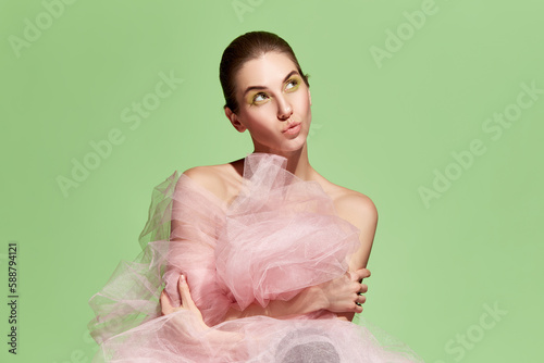 Images of fashionable stylish emotional woman wearing pink tulle dress posing with pensive face over green background. Pink tulle dress