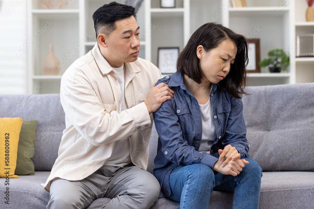 Family psychological support, Asian couple sitting at home on sofa thoughtful, woman comforting and supporting depressed man. Family psychological support, Asian couple sitting at home on sofa thoughtful, woman comforting and supporting depressed man.