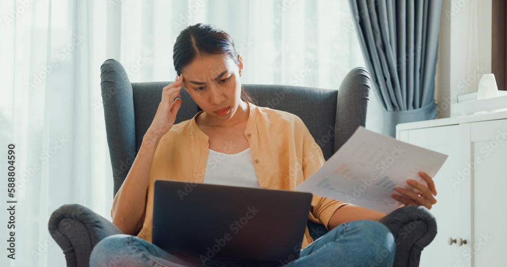 Young attractive asian female girl or university student sit on sofa chair with computer laptop feel focus and serious look on screen and thinking idea work report in living room at cozy home.