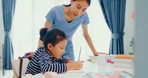 Asian toddler girl with sweater sit in front of desk with notepad use pencil focus on write notebook do math homework with her mother or tutor on weekend at home. Distance online learning concept.