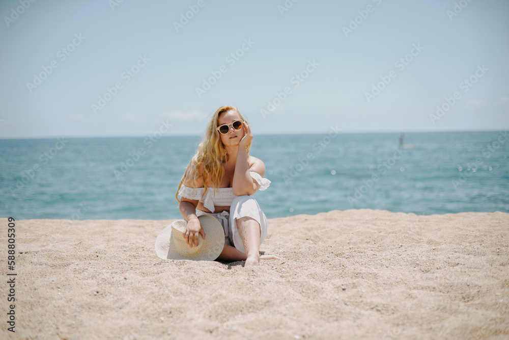 Happy girl in white outfit sits on sand against the backdrop of the sea or ocean beach. Woman smiles and laughs, vacation and joy. Fashion model, beautiful jewelry, earrings, hat, sun glasses Happy girl in white outfit sits on sand against the backdrop of the sea or ocean beach. Woman smiles and laughs, vacation and joy. Fashion model, beautiful jewelry, earrings, hat, sun glasses