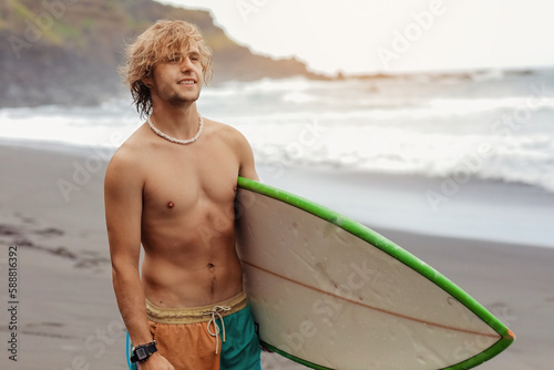 Fit young surfer man with curly blond hair with surfboard goes by the ocean having fun doing extreme water sports, surfing. Travel and healthy lifestyle concept. Sports travel destination.