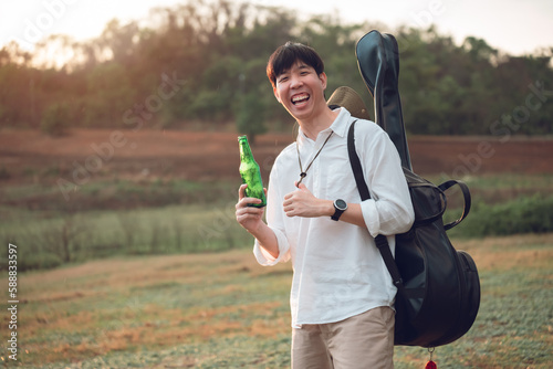 Smiling asian young man holding a bottle of beer and carring guitar at campgrounds, camping outdoors lifestyle on vacation summer.