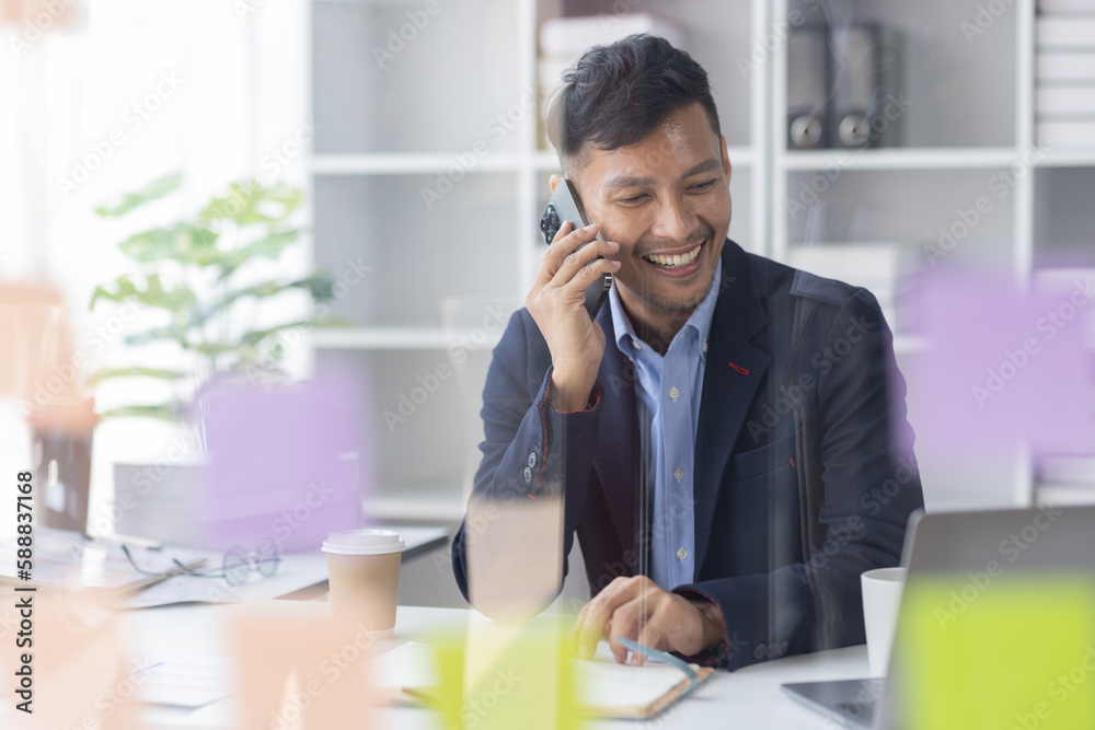 Cheerful business Asian man freelancer making telephone call share good news about project working in office workplace, Asian business man planning finance concept. Cheerful business Asian man freelancer making telephone call share good news about project working in office workplace, Asian business man planning finance concept.