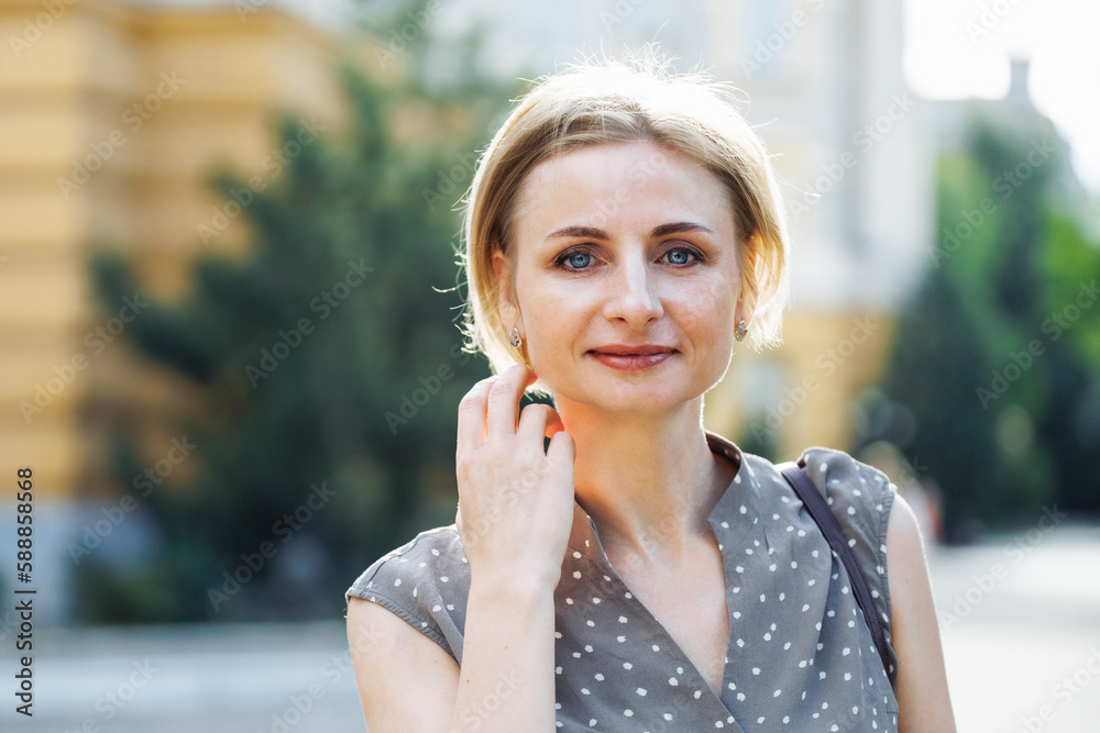 Portrait of gorgeous looking woman standing outdoors among green trees in parkway and touching hair with hand. Middle aged, 40s lady with sky blue eyes. Selective focus, blurred urban background.
