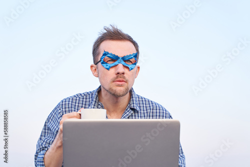 Cheerful man in nightwear shirt and blue face mask using laptop outdoors. IT support superhero guy using laptop holding cup of hot tea or coffee. High quality image © boytsov