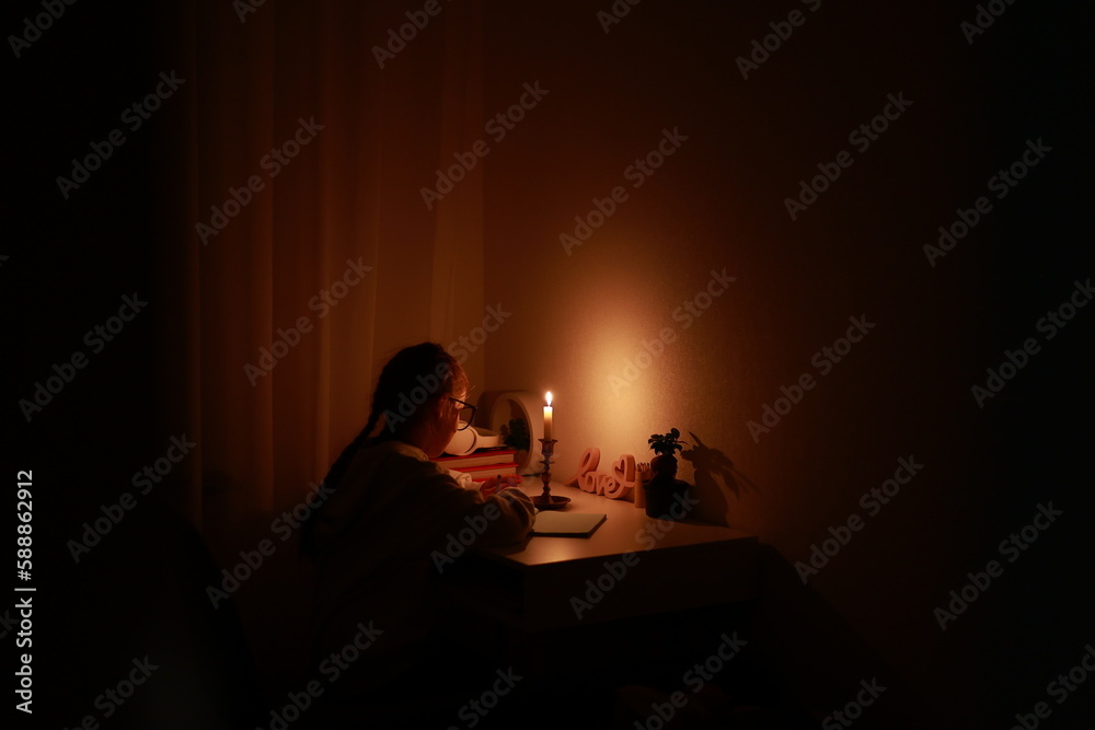Schoolgirl uses candlelight to writes and reads in darkness without electric lights at home. Teen with poor eyesight wears glasses to studies and to do school homework during blackout. Vision problems Schoolgirl uses candlelight to writes and reads in darkness without electric lights at home. Teen with poor eyesight wears glasses to studies and to do school homework during blackout. Vision problems