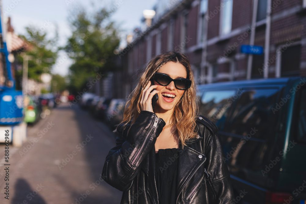 Young cute tourist blonde woman with two thin braids is talking on the phone outside, wearing black leather jacket and black dress, sunglasses. Pretty happy girl with phone in the city.