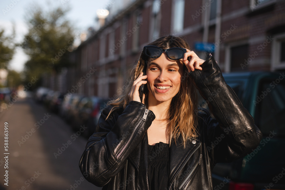 Attractive happy woman talking on cell phone, raised her glasses in surprise, open mouth. Young cute tourist blonde woman with two thin braids outside, wearing black leather jacket and black dress.