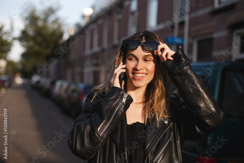 Attractive happy woman talking on cell phone, raised her glasses in surprise, open mouth. Young cute tourist blonde woman with two thin braids outside, wearing black leather jacket and black dress. © zvkate