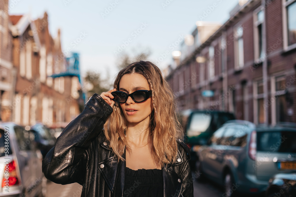 Pensive blonde woman in black leather jacket touch black glasses and posing on street background. Outdoor shot of happy hippie hipster lady with two thin braids and wave hair. Boho freedom style.