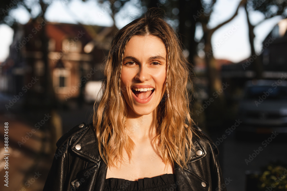 Pensive blonde woman in black leather jacket and dress with open mouth posing on street. Outdoor shot of happy hippie lady with two thin braids and wave hair. Coachella or boho freedom style. - obrazy, fototapety, plakaty 