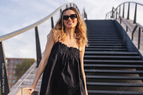 Attractive woman wear black dress and glasses, she have two thin braids and curly hair, girl smiles and walk outdoors standing on the stairs bridge. photo