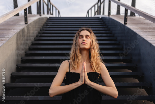 Attractive calm woman with two thin braids and curly hair sitting on the stairs in pray gesture with closed eyes. Girl make thanksgiving symbol on the street. Mindful woman look dreaming.