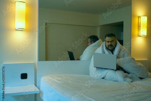 Middle age man using laptop and sitting on bed wrapped in white blanket in his bedroom of hotel. plasma TV hanging on wall. evening light from two lamps.
