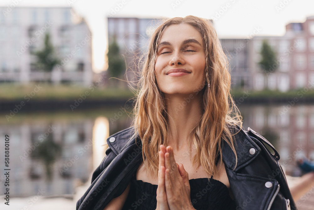 Attractive calm woman with two thin braids and curly hair standing in city in pray gesture with closed eyes. Girl make thanksgiving symbol on the street. Mindful woman look dreaming.