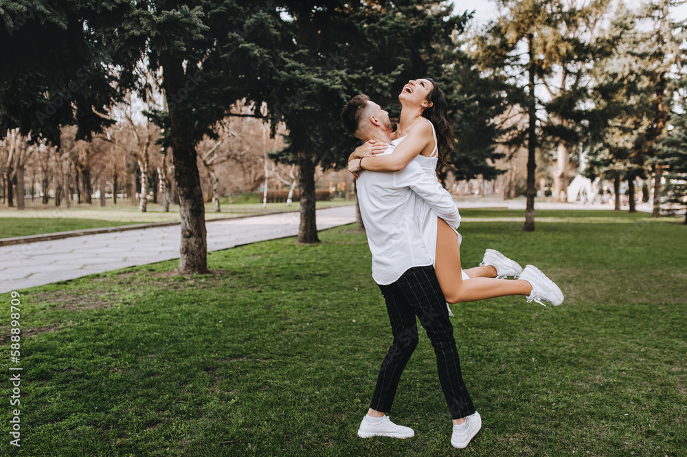 A bearded young groom and a beautiful smiling bride in a white brunette dress are hugging, dancing in a park outdoors. Wedding photography of happy newlyweds, portrait, emotions.