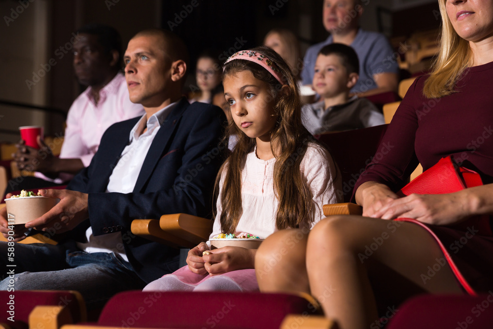 Fototapeta premium Cute preteen girl sitting with her parents in cinema, eating popcorn and watching movie with interest