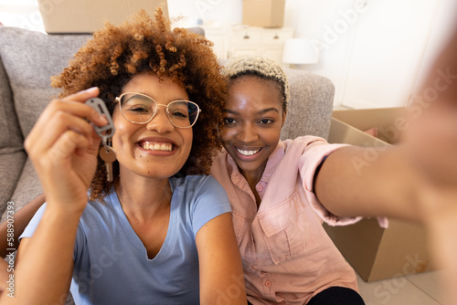 Portrait of cheerful multiracial lesbian couple showing new house key while sitting by sofa at home