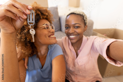 Portrait of smiling multiracial lesbian couple showing new house key while sitting by sofa at home