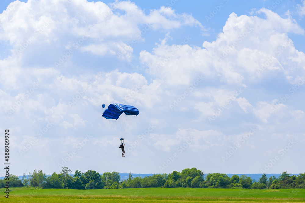 Obraz premium Skydiver with yellow and blue parachute against blue sky and white clouds, landing on field, sports aerial entertainment. Air sports.