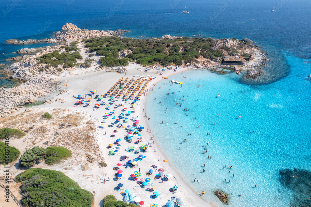Aerial view of colorful umbrellas, sandy beach, swimming people in blue sea, trees at sunset in summer. Sardinia, Italy. Tropical landscape with clear azure water. Travel and vacation. Top drone view