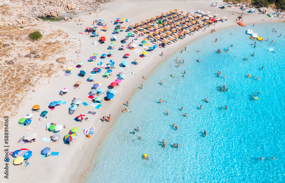 Aerial view of colorful umbrellas on sandy beach, swimming people in blue sea at sunset in summer. Sardinia, Italy. Tropical landscape with clear azure water. Travel and vacation. Top drone view