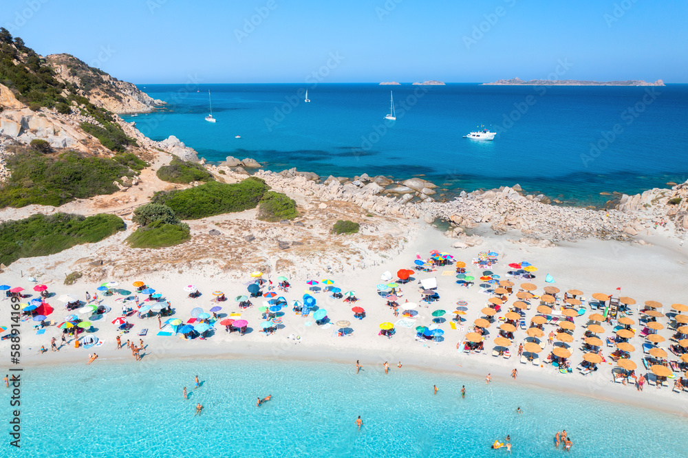 Aerial view of colorful umbrellas, white sandy beach, people, boats and yachts in blue sea, mountain, trees at sunset in summer. Tropical landscape. Travel in Sardinia, Italy. Top drone view