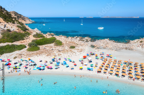 Aerial view of colorful umbrellas, white sandy beach, people, boats and yachts in blue sea, mountain, trees at sunset in summer. Tropical landscape. Travel in Sardinia, Italy. Top drone view